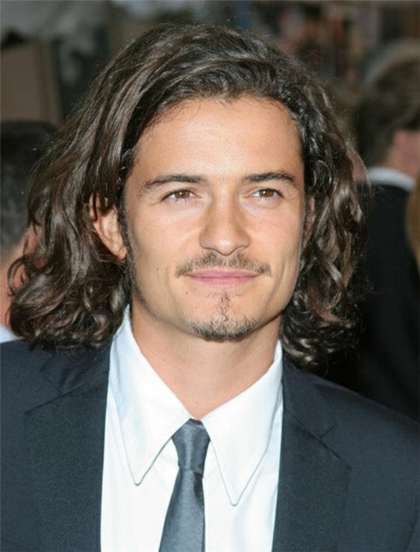 orlando bloom nasmejan, s srednje dolgimi, rjavkasto kodrastimi lasmi, počesanimi na eno stran, kodrastimi pričeskami, kratkimi brki in kozjo bradico, svečano obleko z belo srajco in črno sijočo kravato