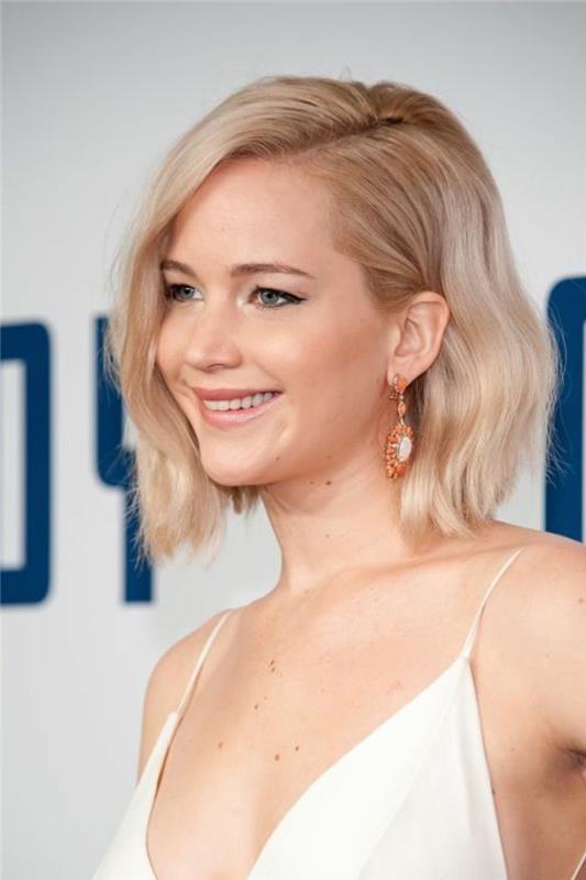 jennifer Lawrence'ın yakın çekimi, dalgalı açık sarı yanları ayrılmış bob'lu, beyaz askılı bluz ve turuncu küpeler takmış.