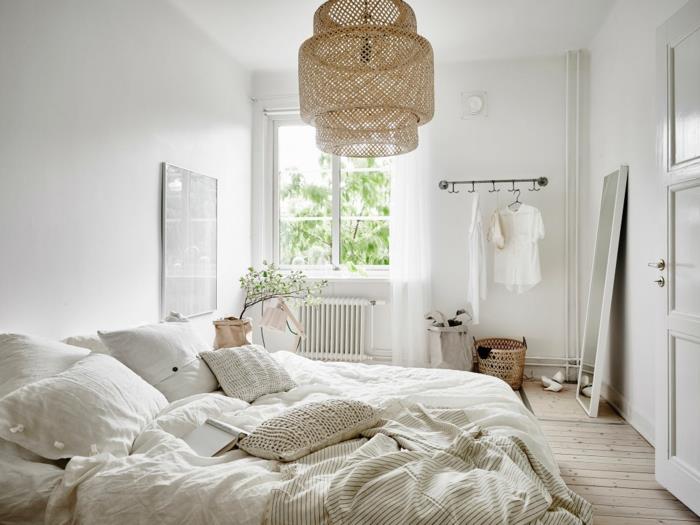 „Cocooning“ dekoras miegamajame, rotango lubų šviestuvas, didelė balta lova su keliomis pagalvėmis, lentos grindys, veidrodis su nugara