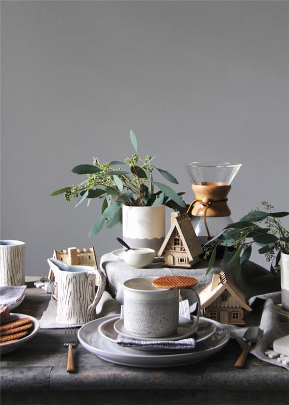 skandinaviško stiliaus kalėdinė dekoracija, kavos puodeliai, arbatinukas, žalios šakelės, šviesiai pilka siena