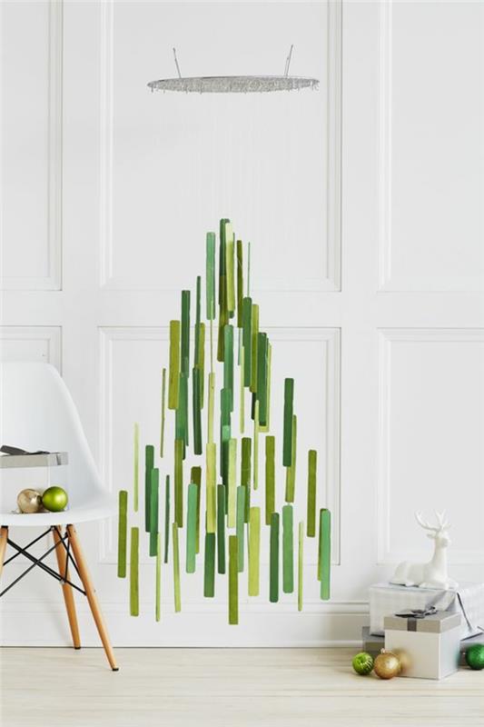 Božična okrasna ročna dejavnost v obliki jelke, kovinski elementi v zeleni barvi različnih dimenzij, postavljeni skupaj postopoma, da tvorijo jelko, beli plastični stol in noge iz svetlega lesa, bele stene in tla, minimalistično vzdušje