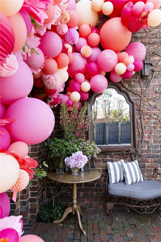 lauko puošmena vakarėliams ir vestuvėms, daugybė rožinių balionų, surištų dekoratyvinėje girliandoje, senas namas, mažas lauko stalas ir kalta ketaus sofa
