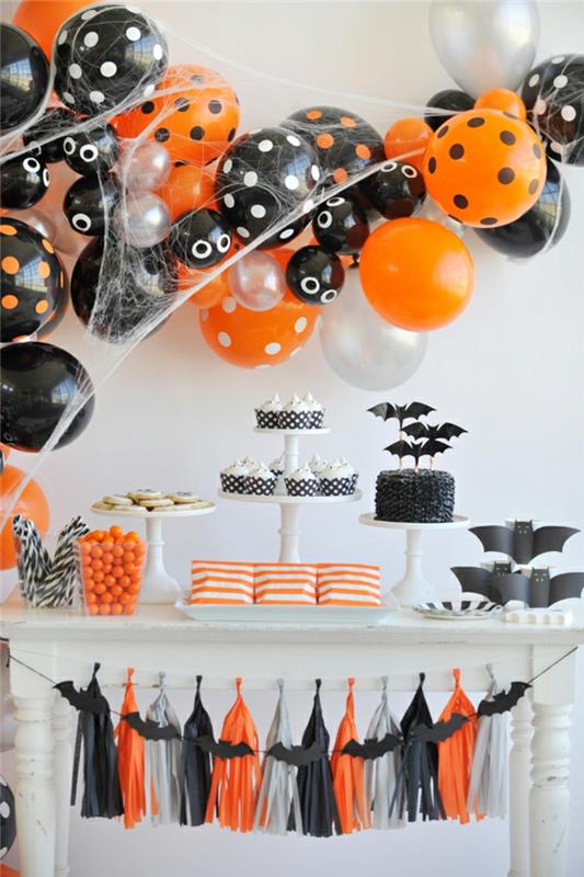 baltas šventinis stalas, Helovino apdaila, pyragai su šikšnosparniais pristatymo padėkluose, oranžiniai saldainiai, dekoratyviniai pakraščiai