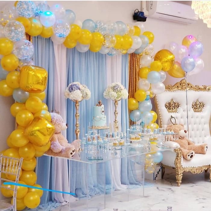akrilo stalas, didelis barokinis fotelis su smėlio spalvos lokiu, geltona ir mėlyna baliono arka, mėlynas pyragas, krištolinė lubų lempa