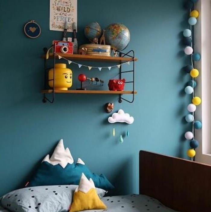 çocuk odasında ördek mavisi dekorasyon, sarı vurgular, top çelenk, sarı ve mavi dağ şeklinde minderler, ahşap ve metal duvar rafı, jiuetler