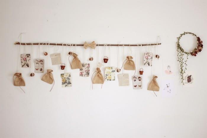 dreifmedžio šaka ant baltos sienos su mažais dovanų maišeliais ir įvairiomis prašmatniomis senovinėmis dekoracijomis, Kalėdų vainikas su džiovintomis gėlėmis, „pasidaryk pats“ advento kalendorius