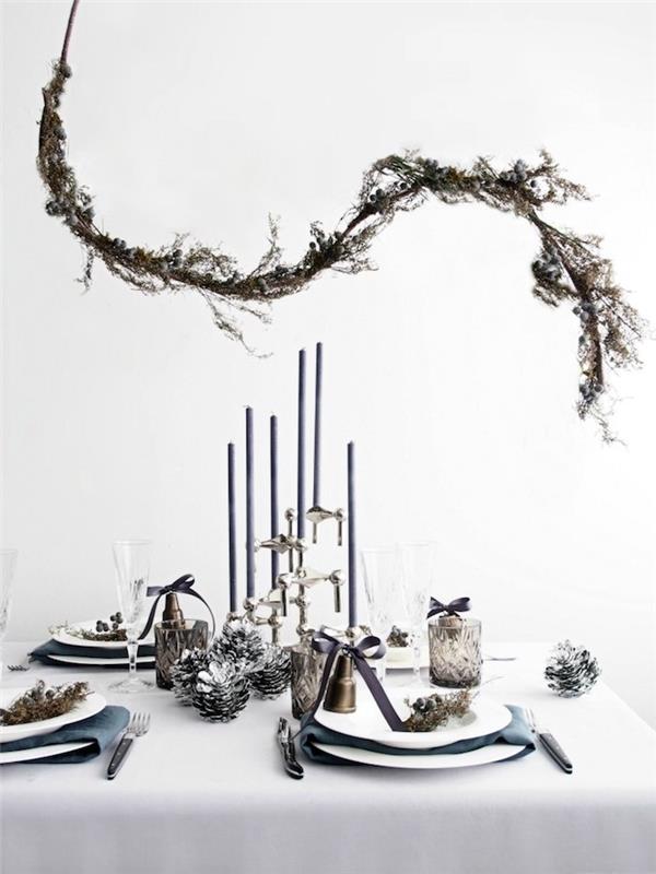 Skandinavska novoletna dekoracija, okrasna veja nad mizo z belim prtom, svečnikom in srebrnimi borovimi storži, vzorci vegetacijske deko