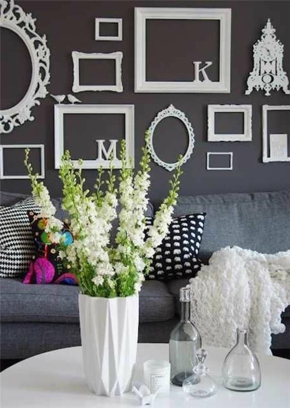 stena iz starih vintage belih okvirjev in okrasnih belih črk, siv kavč, večbarvne blazine, bela vaza, bela miza, šopek rož