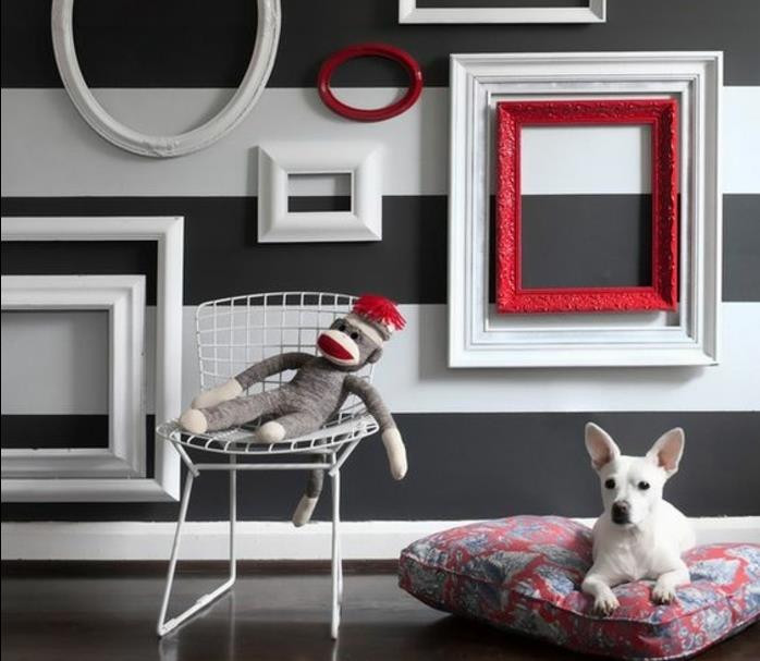 črtasta stena, okrašena s praznimi ovalnimi in pravokotnimi okvirji v beli in rdeči barvi, majhen bel pes na blazini, kovinski stol, opičja igrača, obleči steno