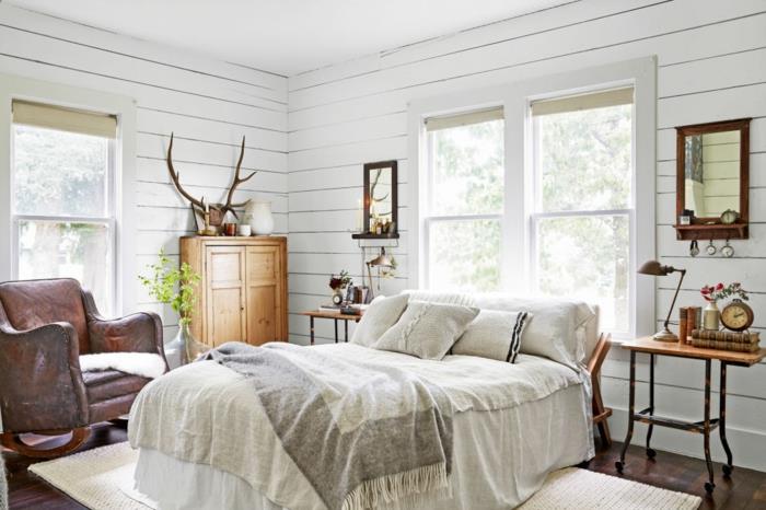 podeželska elegantna spalnica, posteljnina v beli in sivi barvi, rjav usnjen naslanjač, ​​lesena garderobna omara, bela obloga, okras jelenovega roga, vintage nočna omarica in nočna svetilka