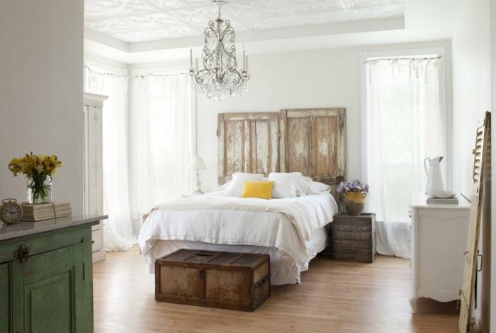 Ideja za dekoracijo podeželske spalnice za odrasle, posteljnina, konec postelje, kovinska skrinja, nočna omarica, leseni zaboj, obrabljena okrasna lesena vrata, baročni lestenec
