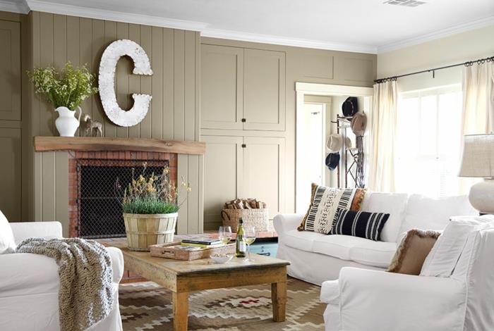 žalio medžio kavos staliukas, balta sofa ir foteliai, dekoratyvinė sieninė raidė, židinys, raštuotas rudas kilimas, įvairiaspalvės pagalvėlės, šiuolaikiškas kaimiškas dekoras