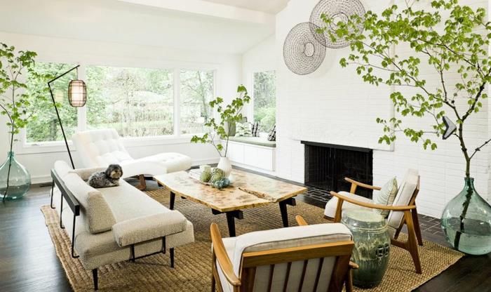 kaimiškas prašmatnus kaimo dekoras, rudas kilimas, balta poilsio kėdė, metalinė sofa su balta sėdynės pagalve, medinės kėdės, dizainerio kavos staliukas, žalių augalų deko, židinys, šuo