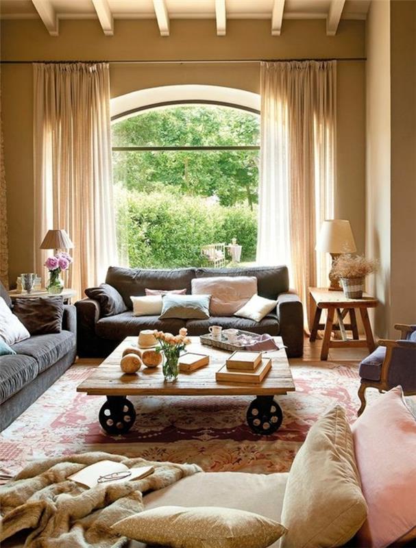 elegantna podeželska dnevna soba, lesena mizica s kolesi, sive zofe, orientalske preproge z vzorcem, blazine v pastelnih tonih, bež stena