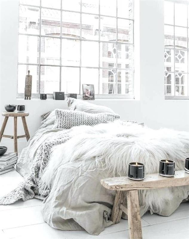 pilkas tinklinis lovos užtiesalas, neapdorotas medinis lovos galas Šiaurės Skandinaviško stiliaus miegamajame, naktinis stalas, taburetė ir aromatinės žvakidės, romantiškas palėpės deko