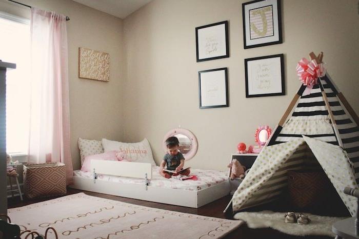 kūdikio mergaitės miegamojo dekoravimo idėjos montessori stiliumi su smėlio spalvos sienomis, juodos ir baltos spalvos mergina su juostelėmis, balta lova ant grindų, dekoratyviniai rėmai