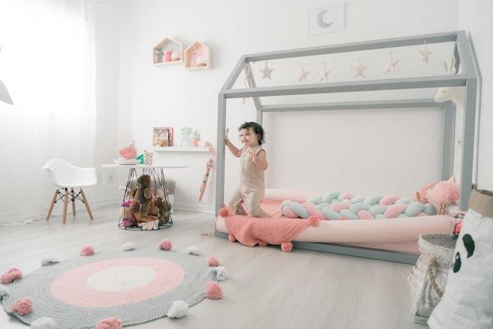 pilkos „Montessori“ kabinos lovos su rožiniu čiužiniu, pilkos ir rožinės spalvos kilimėlio, šviesaus parketo, stalo ir žemos kėdės pavyzdys, įdomios lentynos