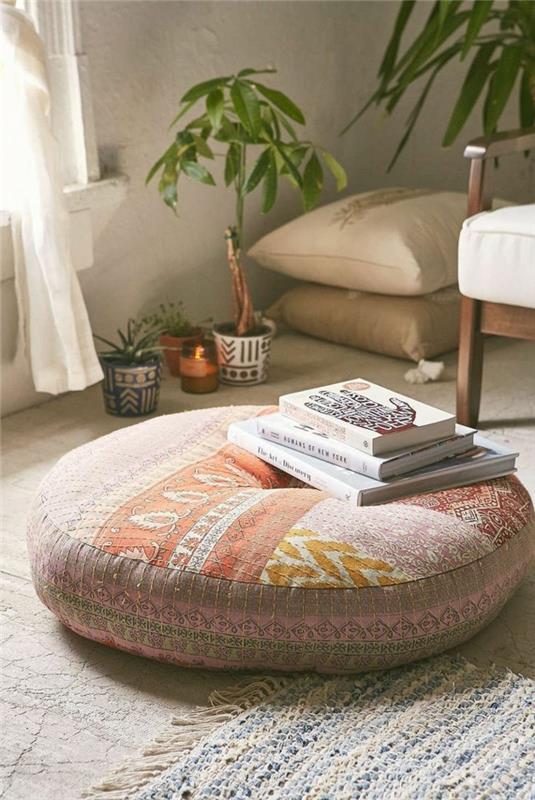 bohemiška svetainė, smėlio spalvos taburetė, knygos, žali augalai, kilimas su pakraščiais