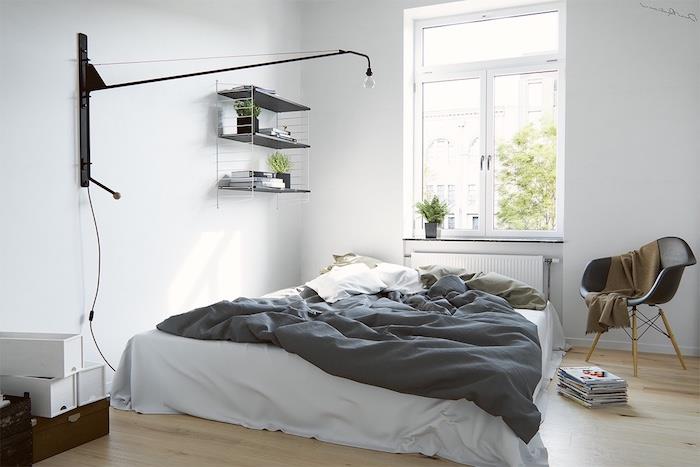 dekoriranje spalnice, postelja na tleh, sivo -belo posteljnina, črni skandinavski stol z rjavo odejo, siva stenska polica