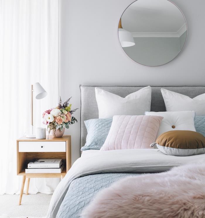 dekoriranje spalnice v pastelnih tonih, siva, bela in praškasto roza posteljnina, miza, lesena nočna omara, šopek svežega cvetja, okroglo ogledalo