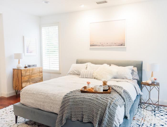 sivo -bela spalnica, bela barva sten in belo perilo na sivi postelji, preproga z geometrijskim vzorcem, lesena komoda, rjav parket in kovinska nočna omarica