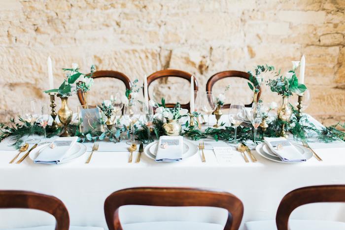 vestuvių stalo dekoras, balta staltiesė, žalumynų centras, gėlės mažose auksinėse vazose, baltos žvakės, akmeninė fono siena, priėmimas tvarte