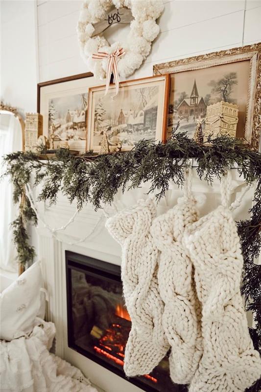 Yeşillikler içinde çelenkler, beyaz örgü çoraplar ve çerçeveli resimlerle Noel şöminesi dekorasyonu