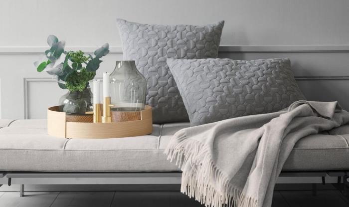 būtini skandinaviško „hygge deco“ gyvenamojo kambario priedai, pilkos pagalvėlės su tekstūruotais raštais, jauki antklodė ir gražus dekoratyvus medinis padėklas su maža keramine vaza