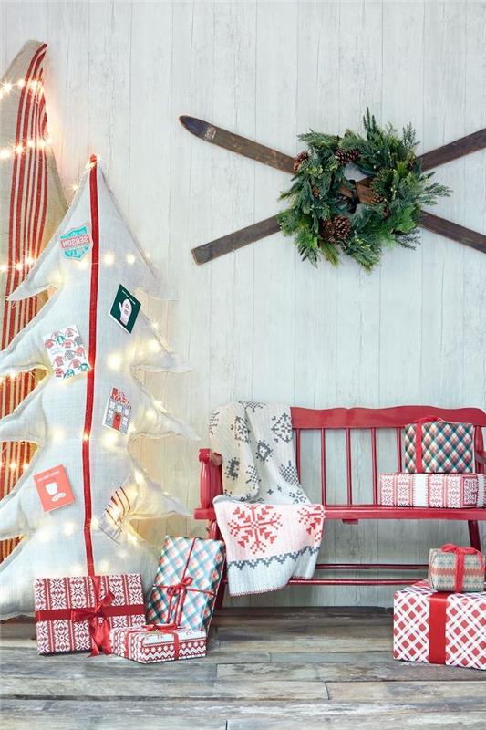 Kalėdinė puošmena su kartonine dovanų eglute ir ant sienos kabančiu raudonu slidžių suoleliu