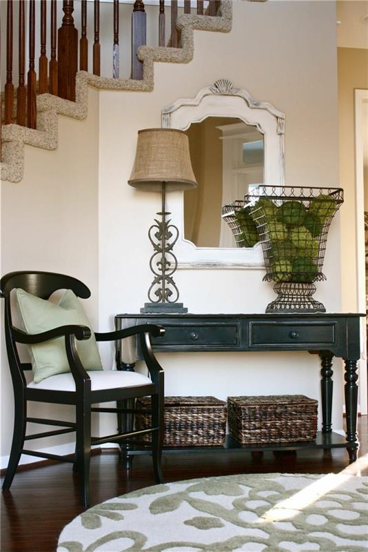 sukurkite prašmatnų įėjimo dekorą su nedideliu juodu stalu, kėdute, apvaliu kilimėliu ir barokiniu veidrodžiu