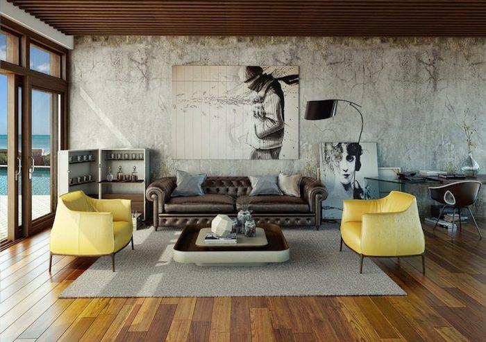 industrijska dekoracija dnevne sobe s temno rjavim usnjenim kavčem, rumenimi naslanjači, sivo preprogo, rjavim parketom, obrabljeno steno, lesenim stropom, črno -belimi grafičnimi okvirji