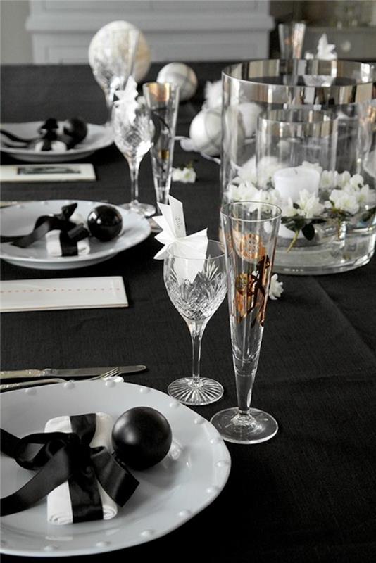 Novoletna dekoracija v črni barvi z belimi krožniki in kristalnimi kozarci šopek na sredini mize