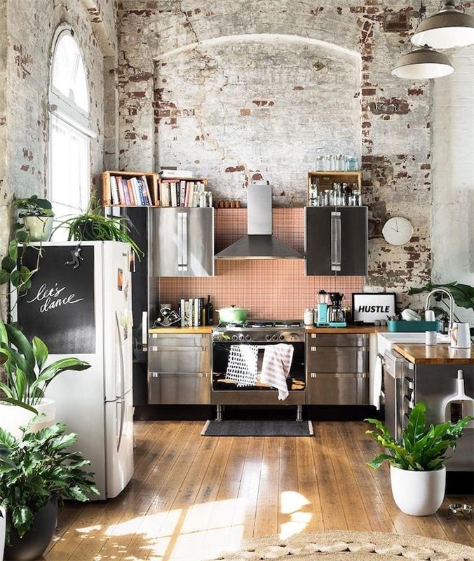 ideja o industrijski kuhinjski dekoraciji z opečnimi stenami, kuhinjsko fasado iz nerjavečega jekla, belim hladilnikom s tablo, surovim parketom in vzorcem džungle, predstavljenim z rastlinami