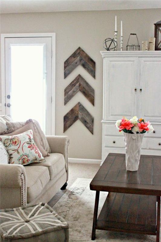 prašmatnus kaimo dekoras, medinis kavos staliukas, pilka sofa, gėlių rašto pagalvėlės, pilka kėdė, patinuota medinė indauja, medinių strėlių sienų dekoras