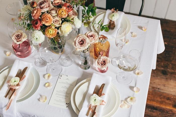 poceni poročni dekor, rože, razporejene v krmilih, pisane steklenice in kozarci ter cvetni listi vrtnic, raztreseni po mizi, beli prtički in jedilni pribor, okrašeni s cvetjem in rožnatim trakom