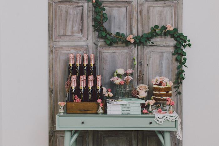 vintage poročni deco, ozadje surovih lesenih vrat z okrasjem cvetličnega venca, pastelno zelena miza s steklenicami šampanjca, torta, knjige in cvetlični okras