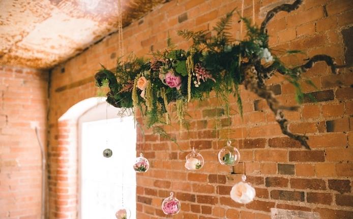 kaimiškos prašmatnios vestuvės, lapija ir gėlėmis dekoruota medinė šaka su mažais kabančiais terariumais su gėlėmis, mūrinės sienos fonas