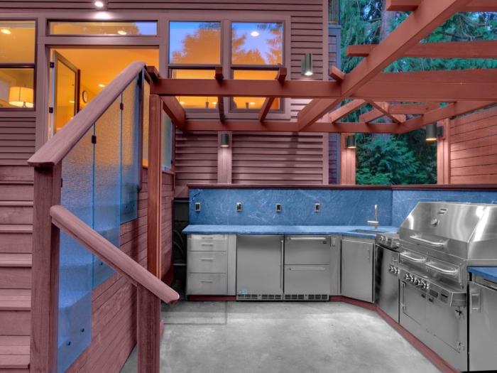 virtuvės konstrukcija sode su mediniu stogu ir nerūdijančio plieno moduliais bei mėlynomis plytelėmis, L formos virtuvės modelis