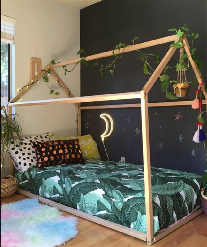 Montessori pedagogika, kūdikio kambario išdėstymas, medinė žaidimų namelio lova, čiužinys, įvairiaspalvės pagalvės, juoda akcento siena, kelių atspalvių kilimas, šviesi girlianda