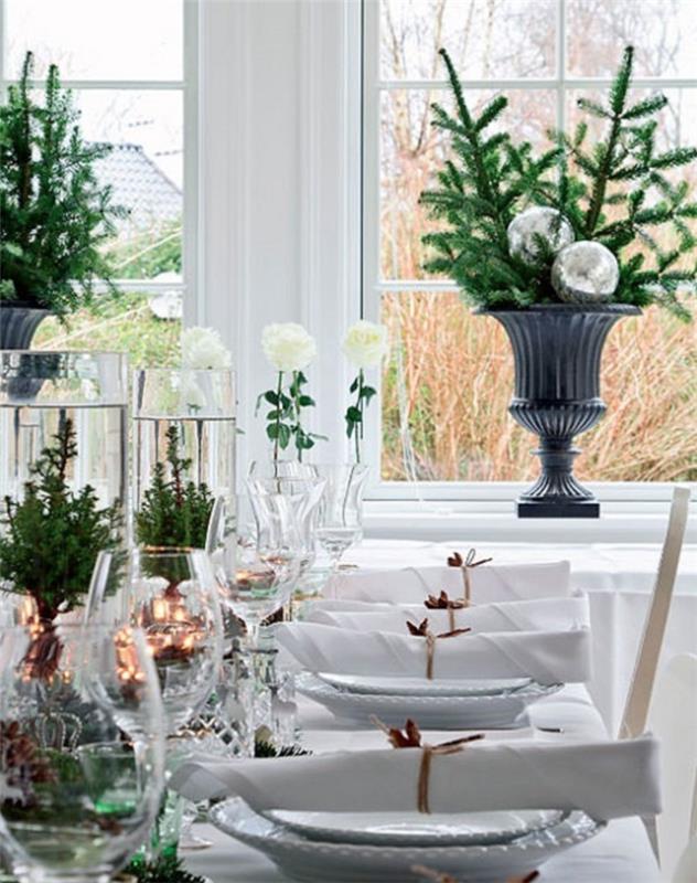 baltas šventinis stalas, baltos lėkštės, didelės tamsiai pilkos vazos su pušies šakomis ir dekoratyviniais kamuoliukais, didelės vyno taurės