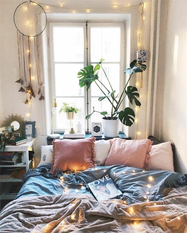 gražus miegamojo išdėstymas, rožinės pagalvėlės, pilka patalynė, baltas vazonas su žaliu augalu, langas, svajonių gaudyklė, šviesi girlianda