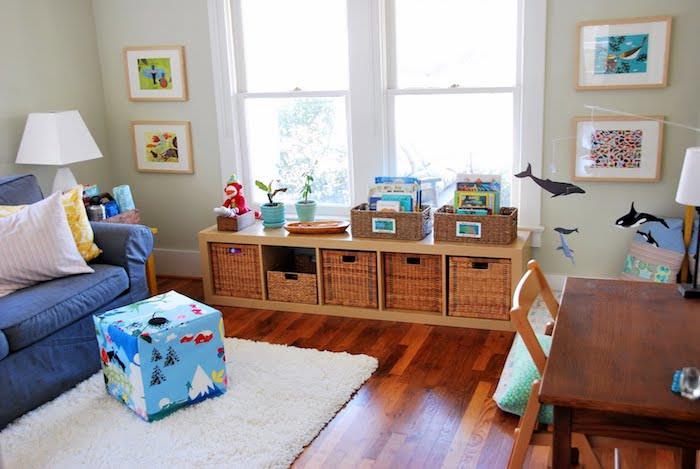 Žema medinė lentyna su sandėliuku žaislų krepšeliuose, rudas parketas, baltas kilimas, mėlyna sofa, aktyvus čiužinys ant grindų, montessori kampelis svetainėje