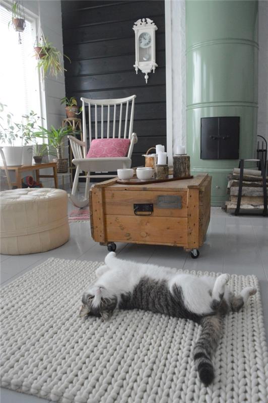 židinio kampas „hygge deco spirit“ su regeneruoto medžio kavos staliuku, supama kėdė ir odinis pufas, miela katė, gulinti ant pilko megzto kilimėlio priešais židinį