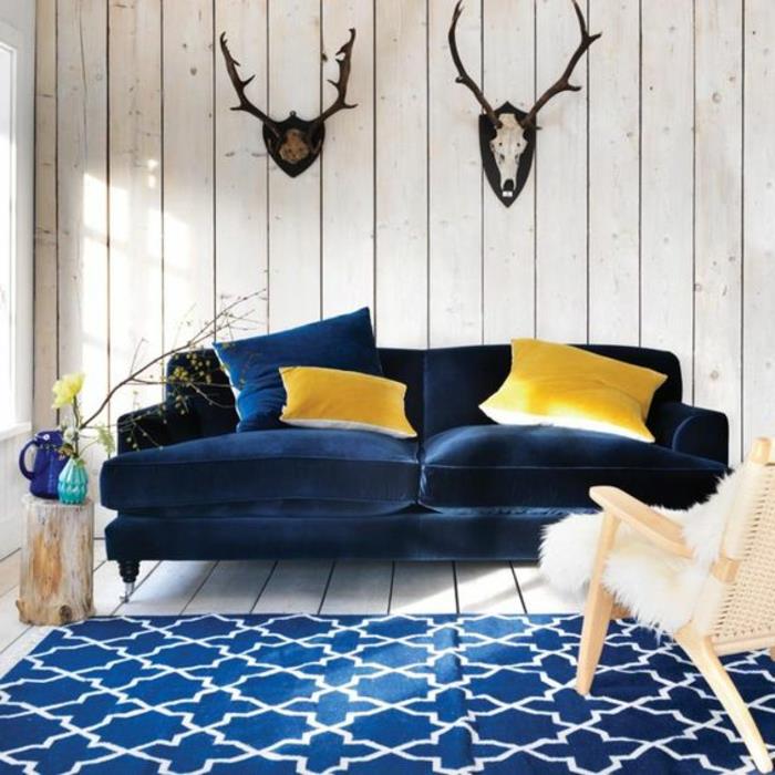 İskandinav oturma odası dekoru, lacivert kanepe ve halı, ahşap paneller, avcılık kupaları, kürklü İskandinav sandalyesi, dekoratif ahşap kütük, sarı minderler