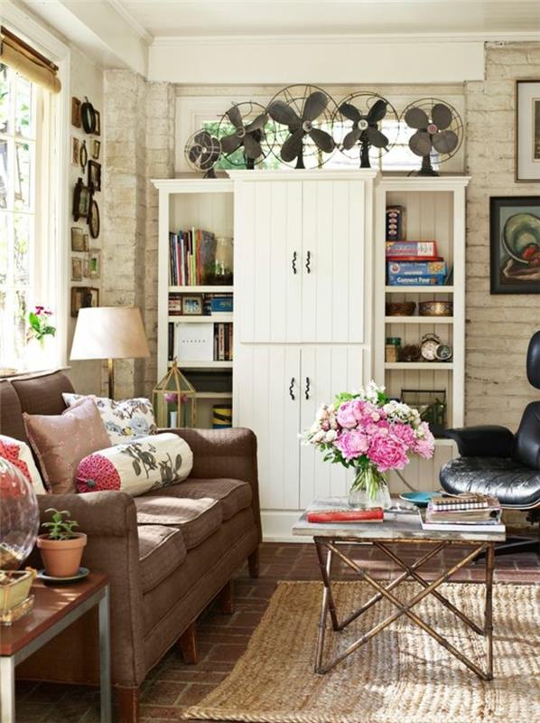 elegantna podeželska dnevna soba, knjižna omara z omarami, rjav kavč, mizica, kamnita stena, okrasni okvirji, šopek rož