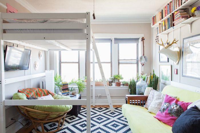 aukšta lova su sukietėjusiu foteliu, pridengtu dessiju, juodai baltas kilimas, medinė ir žalia sofa, baltos sienos, žali augalai ir aukštos lentynos
