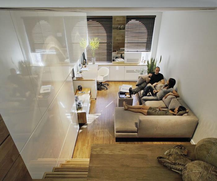 kuhinja odprta za manjši dnevni prostor s sivim kavčem, bela kuhinja, parket iz svetlega lesa, visoka medetažna postelja