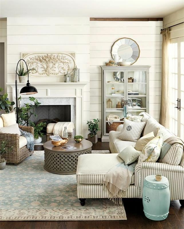 kaimiško stiliaus deko, gėlių rašto kilimas, dryžuota sofa ir rotango fotelis, apvalus rytietiško stiliaus kavos staliukas, židinys, senovinė balta indauja, rudas parketas, prašmatnus kaimiškas dekoras