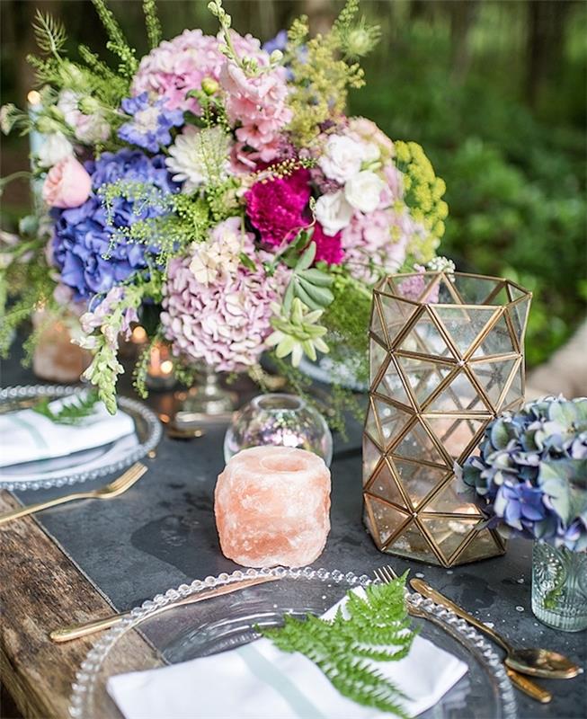kaimiškas vestuvių stalo dekoras su spalvingomis gėlėmis, rožine, fuksija ir violetinėmis spalvomis, žvakėmis, stiklo lėkštėmis ir žaliais lapais servetėlėje