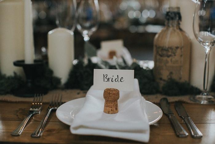 dekoracija poročne mize, beli prtiček, pluta z kartonom z imenom gosta, kmečka lesena miza, sveče, steklenica, venček deco
